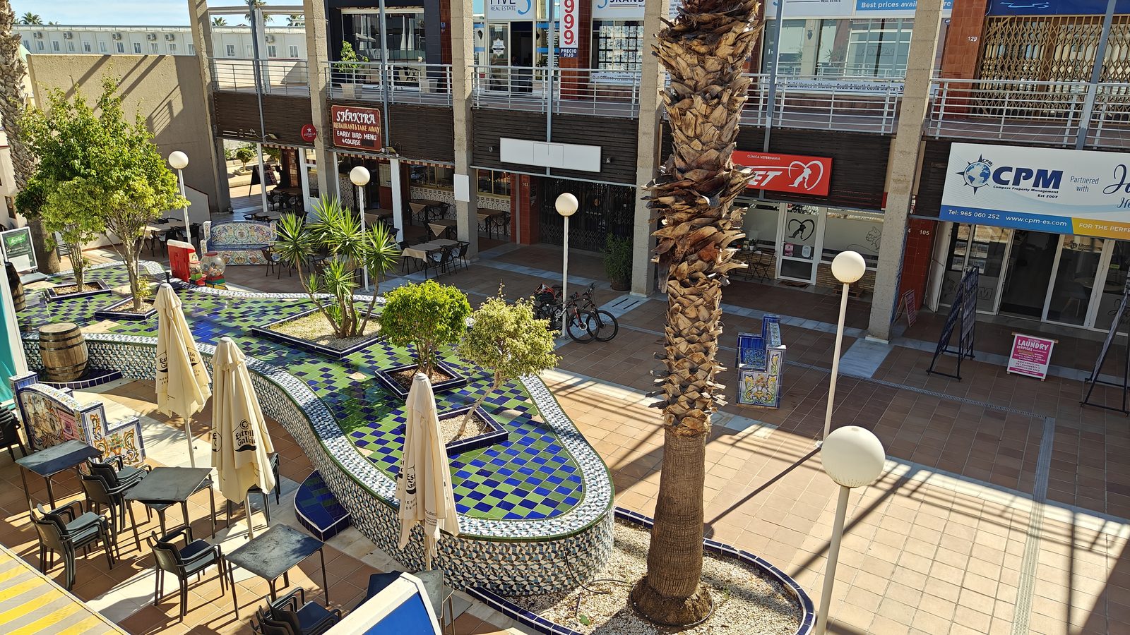 Open-air plaza with a tiled fountain, café terraces and palm trees in Los Dolses' shopping and dining area